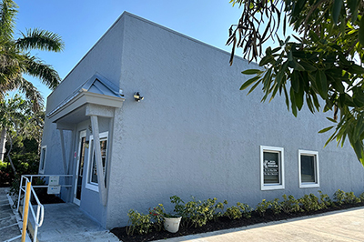 The image shows a single-story building with a flat roof, painted in light blue. There is a small entrance on the left side of the building, and it appears to be a commercial or public space given the architectural style and presence of landscaping around the entrance area.