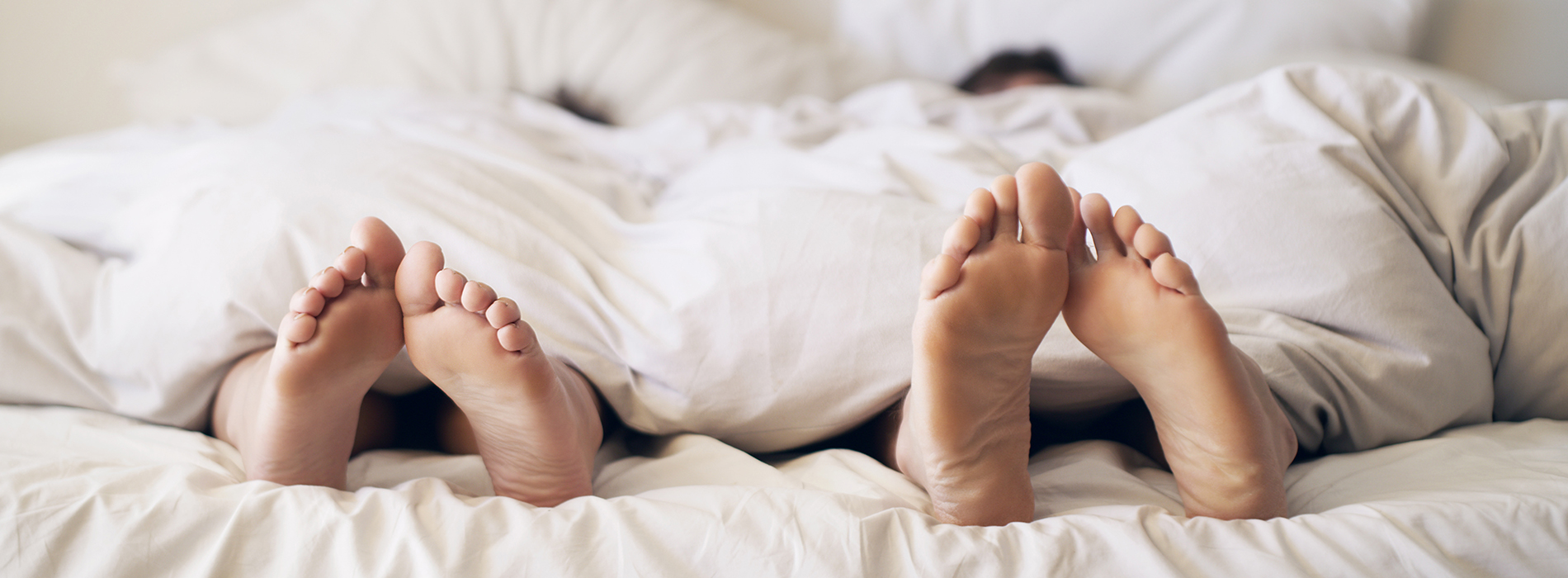The image depicts a pair of bare feet resting on a bed with white sheets.